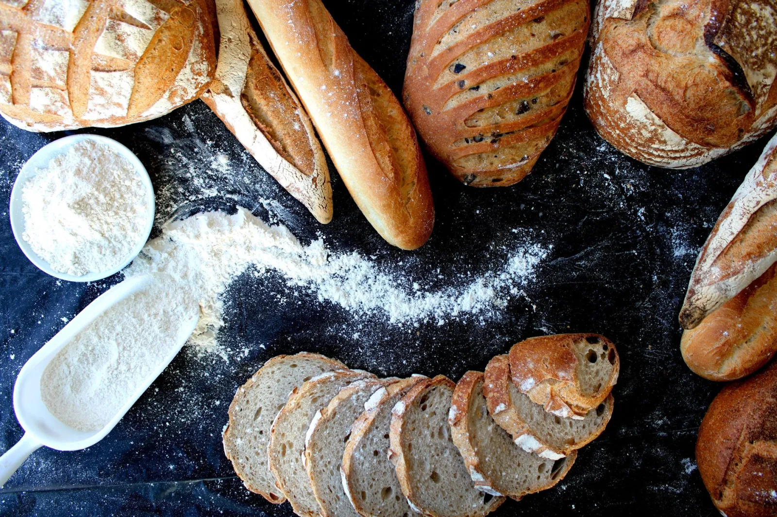 Top view of various freshly baked artisan breads and flour on a dark surface, showcasing texture.