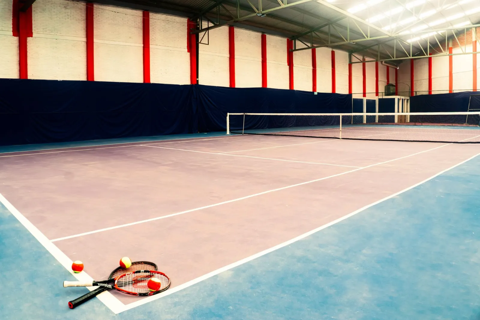 Empty indoor tennis court with rackets and balls in the foreground, showcasing a sports arena setup.