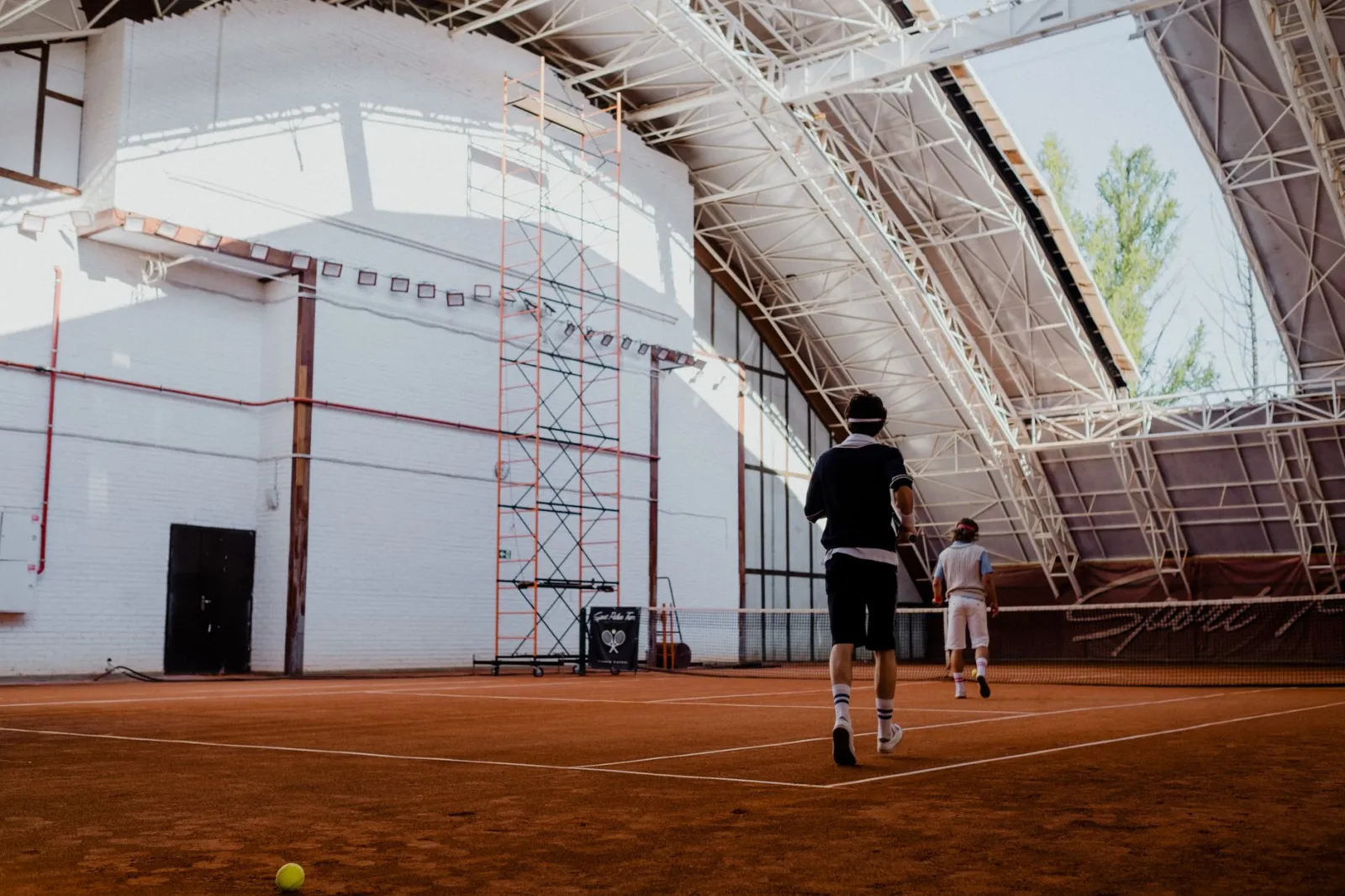 Two players engaging in a tennis match on an indoor clay court with natural lighting.